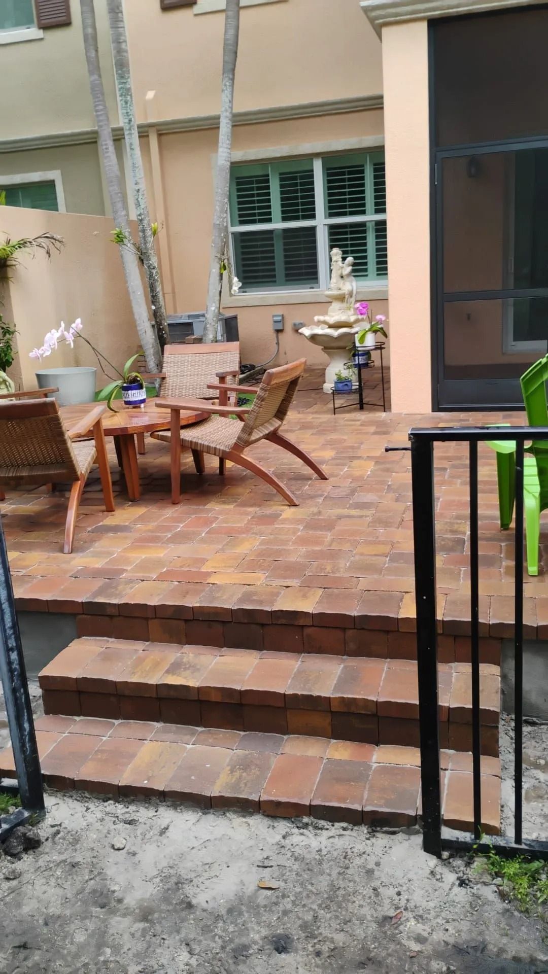 A patio with brick pavers, two wooden chairs, a small table, a stone fountain, and steps leading to a yard.