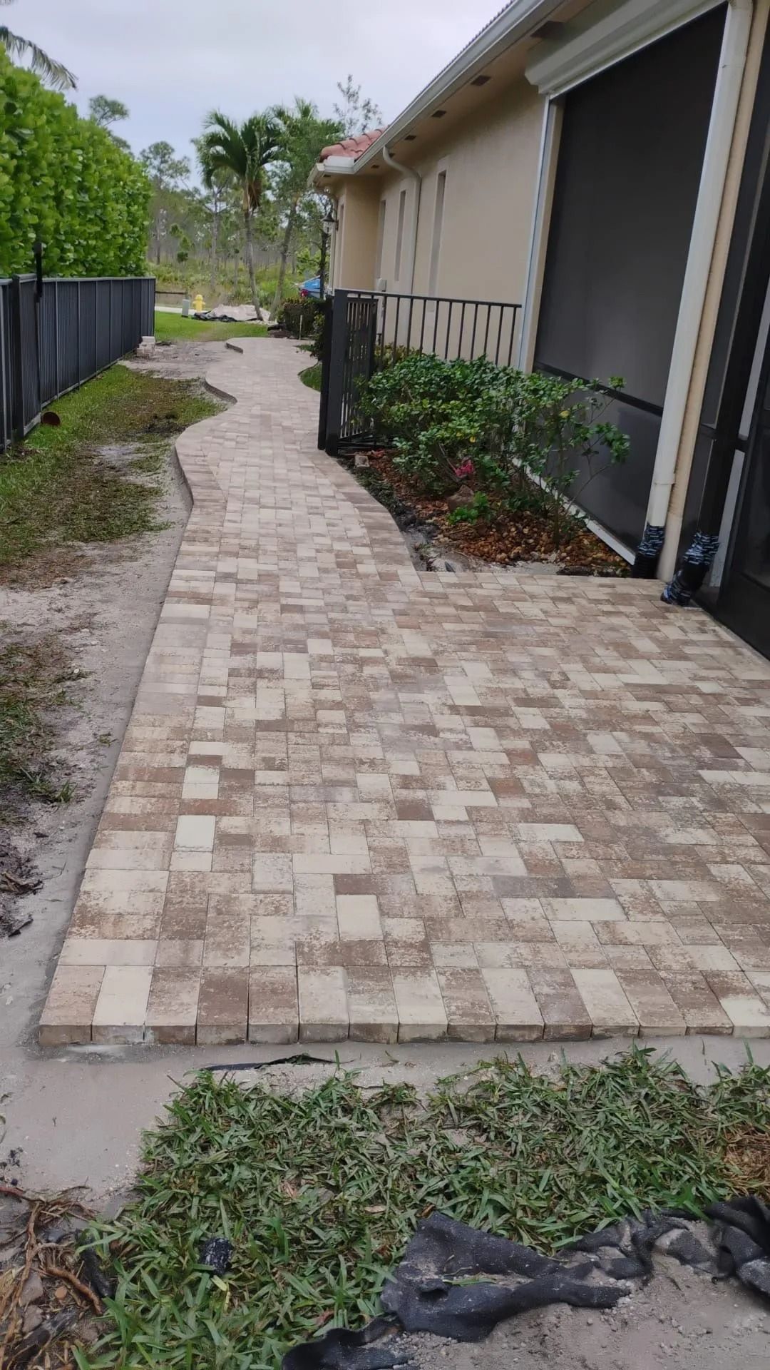 A tan brick paver walkway extends along the side of a house toward a green backyard with palm trees.