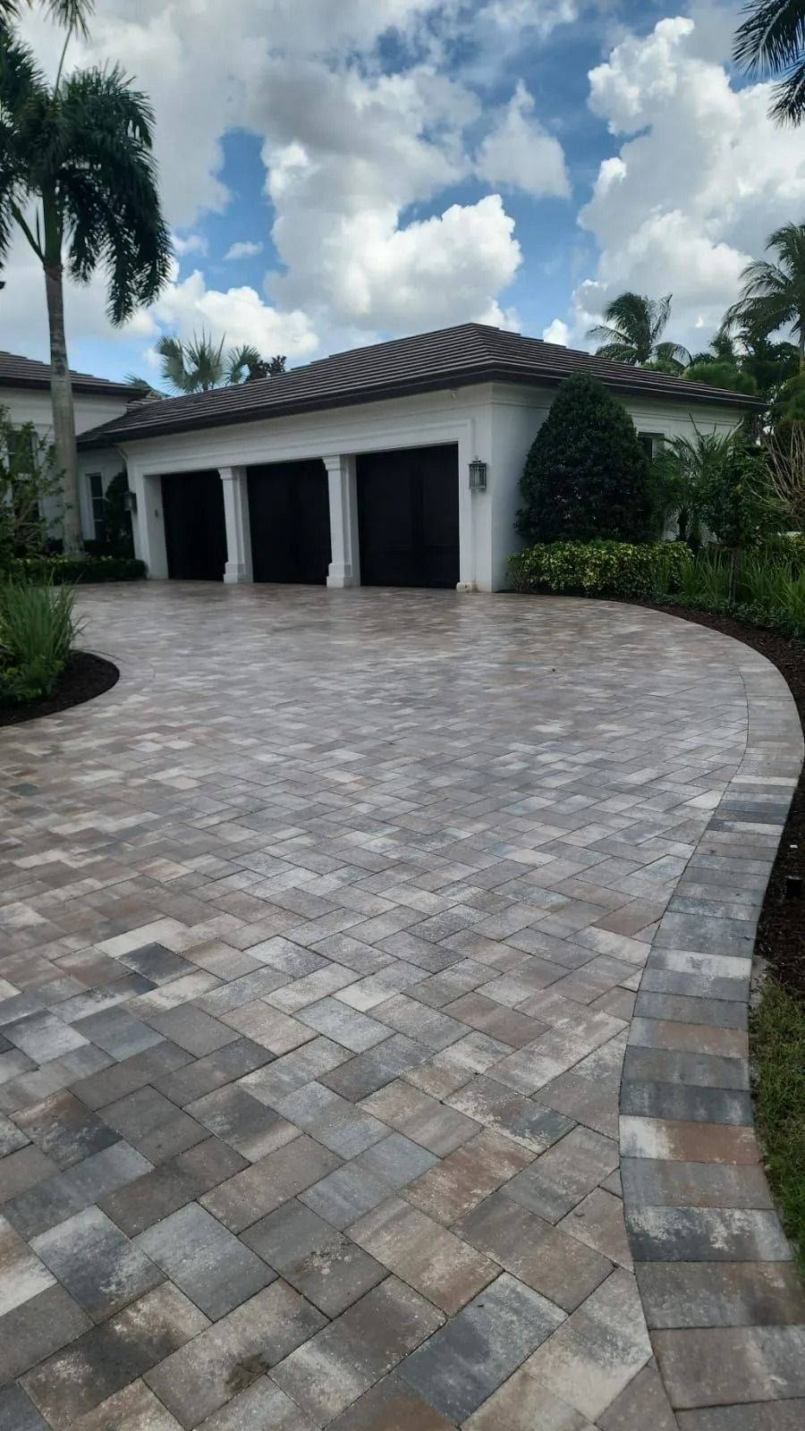 A three-car garage with black doors and a tiled roof, fronted by a curved paver driveway on a sunny, partly cloudy day.