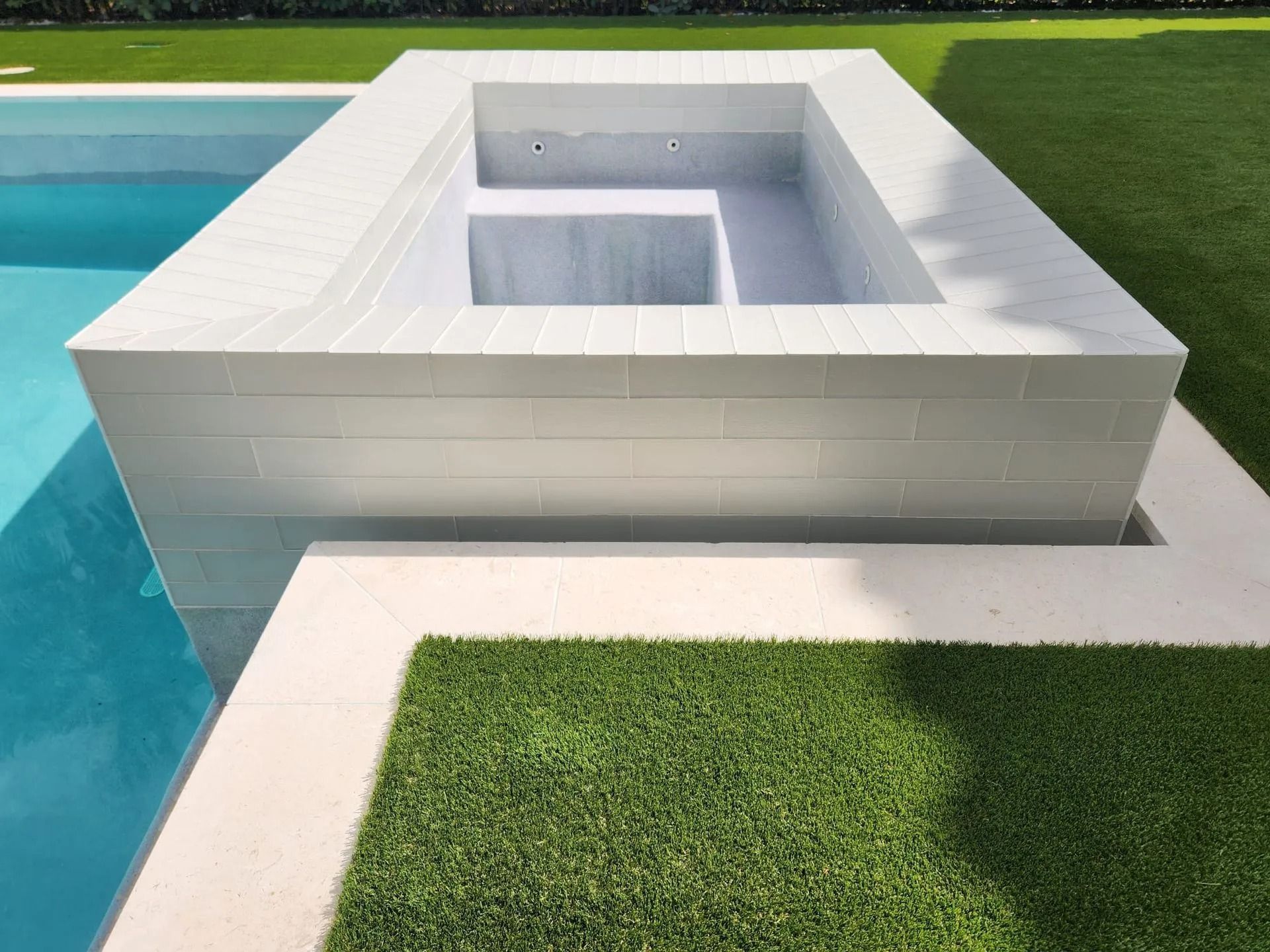 A raised, rectangular white-tiled hot tub next to a swimming pool, bordered by green artificial grass.