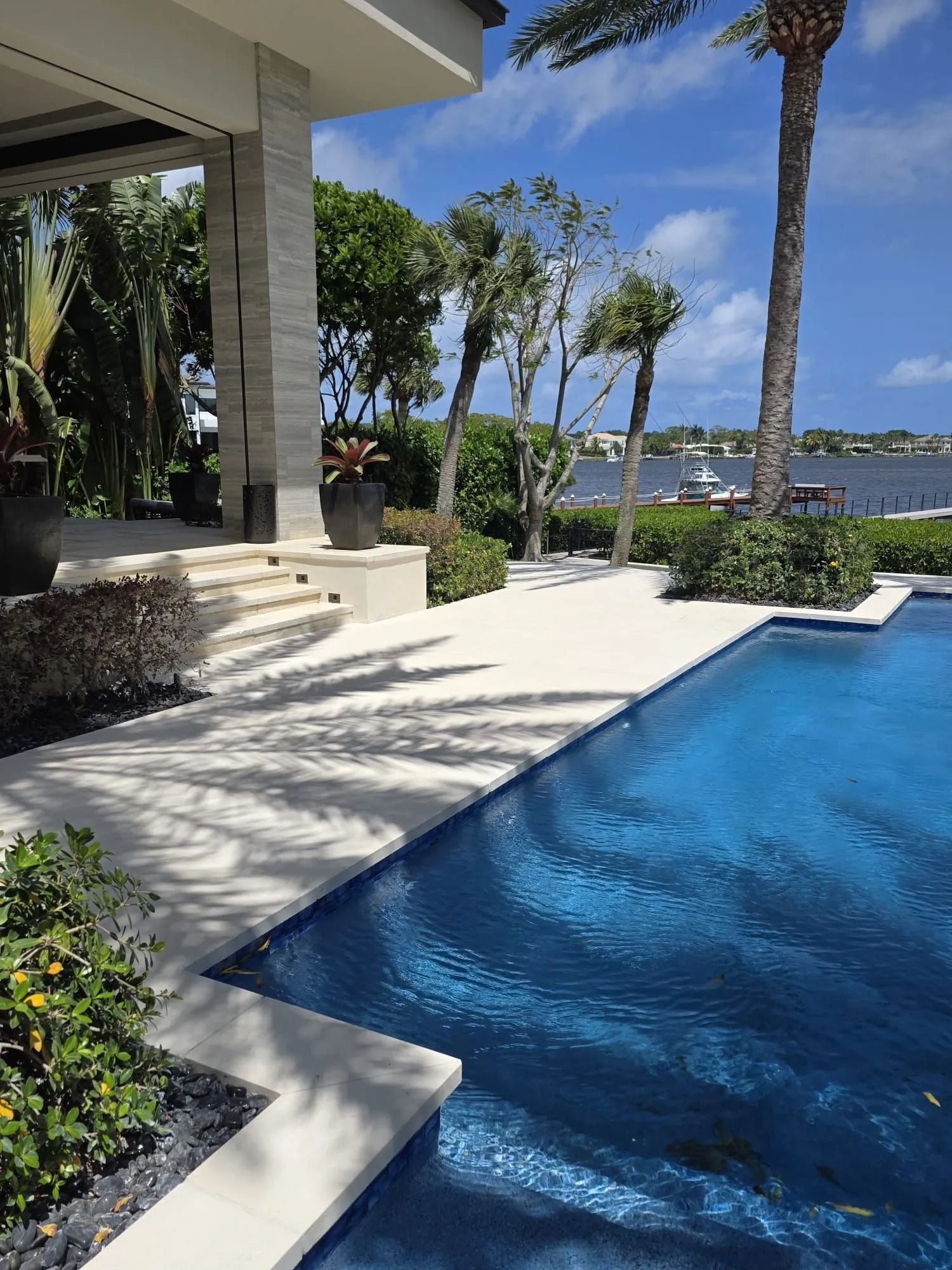 A bright blue pool beside a stone patio and modern home, with lush palm trees and a view of a calm waterway.