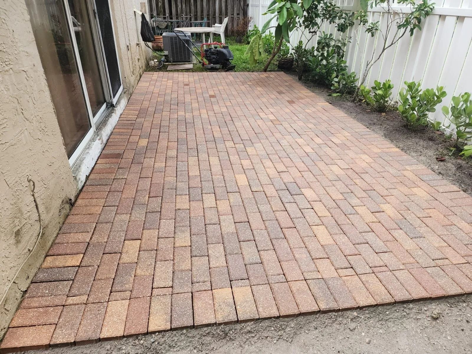 A rectangular patio made of reddish-brown bricks in a herringbone pattern, located beside a house with a sliding door.