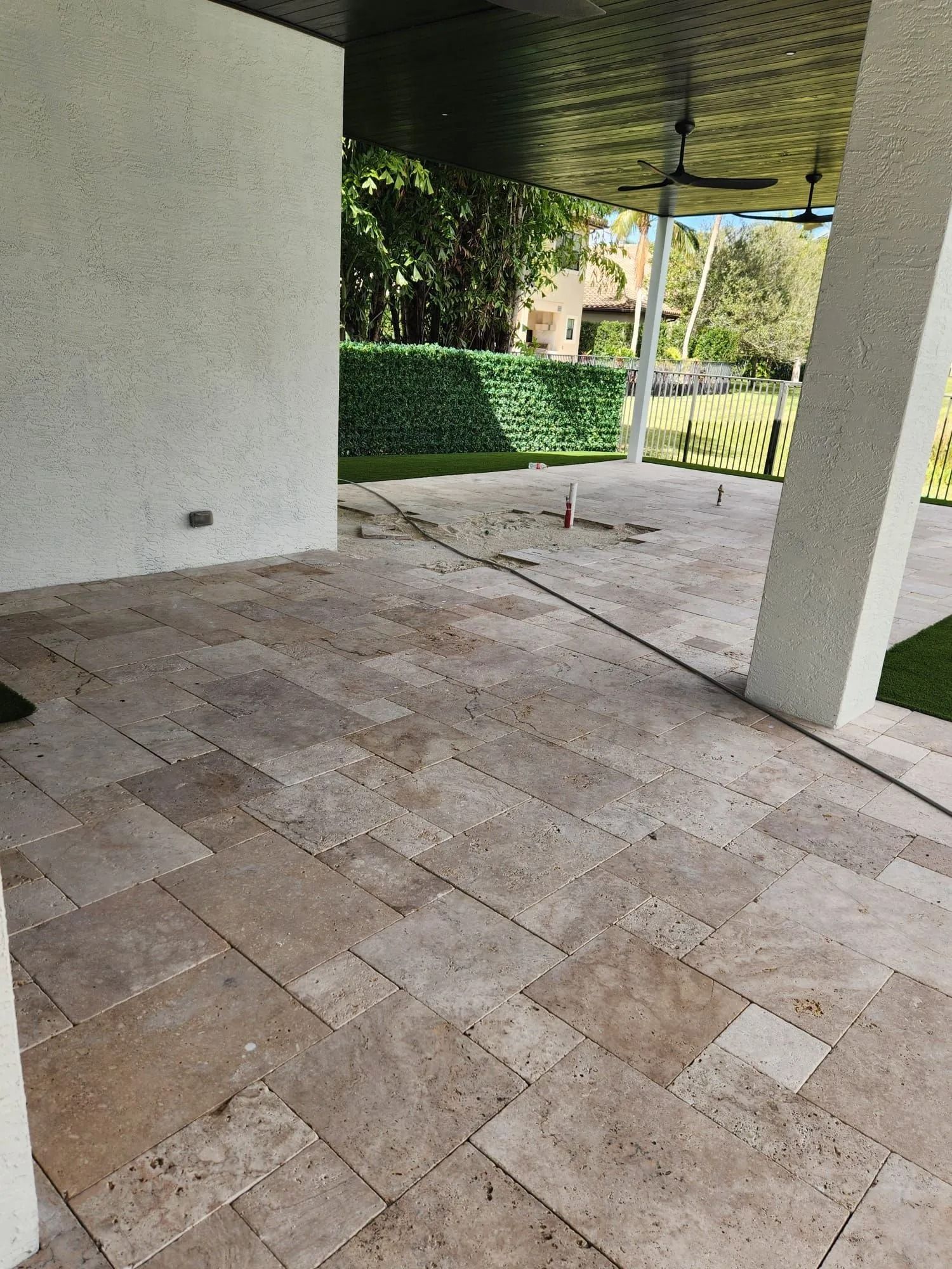 A covered patio with light-colored stone pavers in a patterned layout, leading toward a green hedge and outdoor greenery.