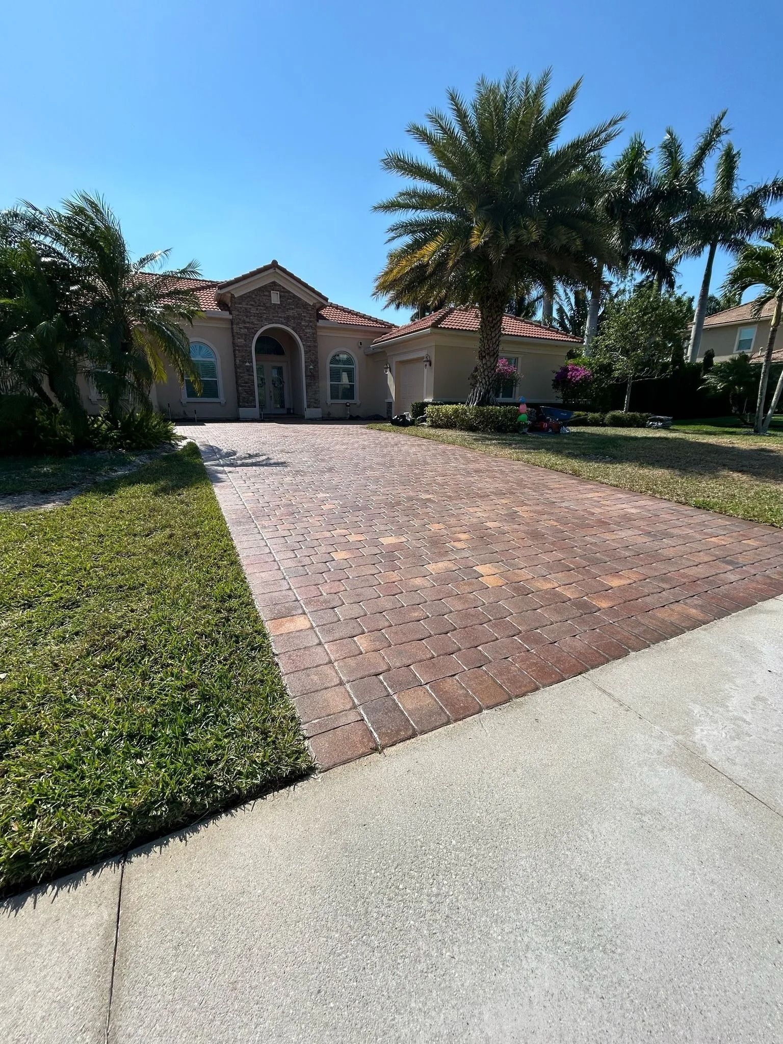 A single-story home with a tan stucco exterior, tile roof, and a paved brick driveway under a bright blue sky.