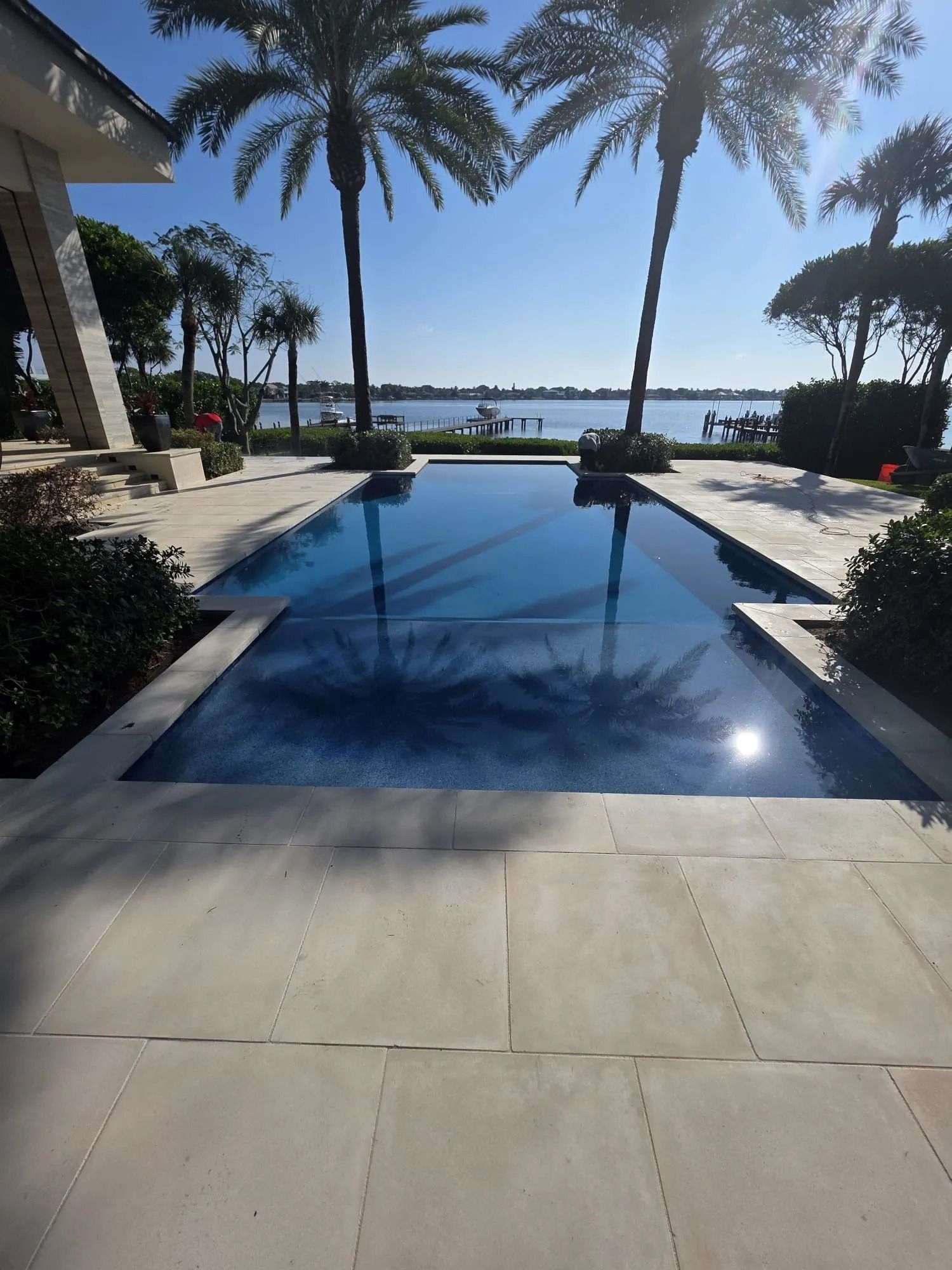 A long rectangular swimming pool stretches toward a scenic lake lined with palm trees on a sunny day.