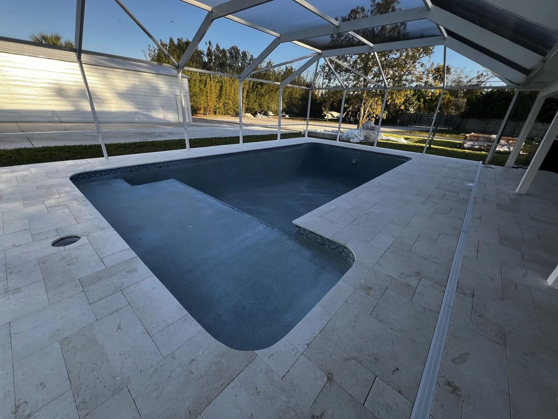 A rectangular dark blue swimming pool with a step ledge, surrounded by light stone pavers inside a screened-in enclosure.