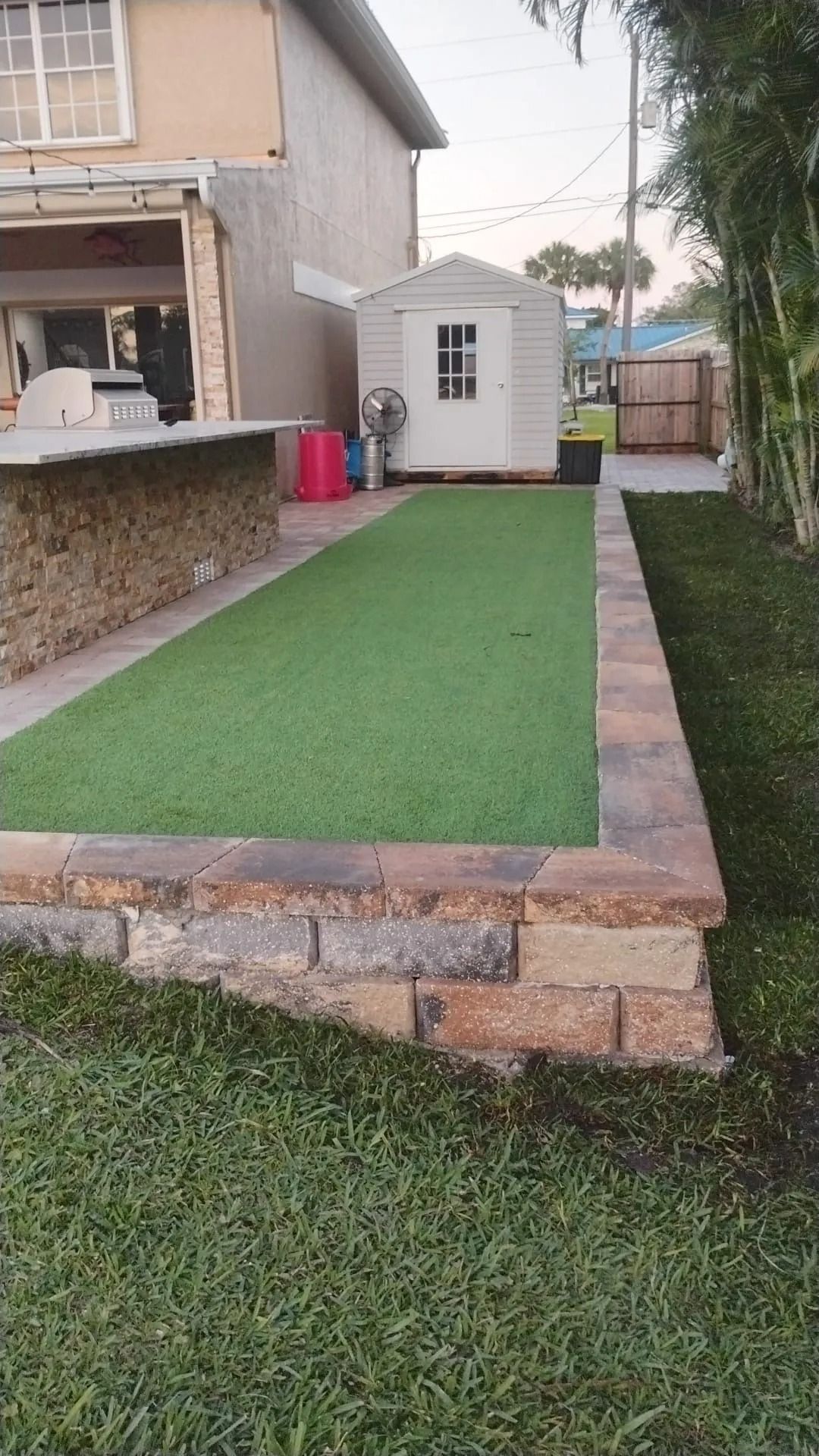 A raised patio area featuring green turf, surrounded by a stone block border, next to a house and a small storage shed.