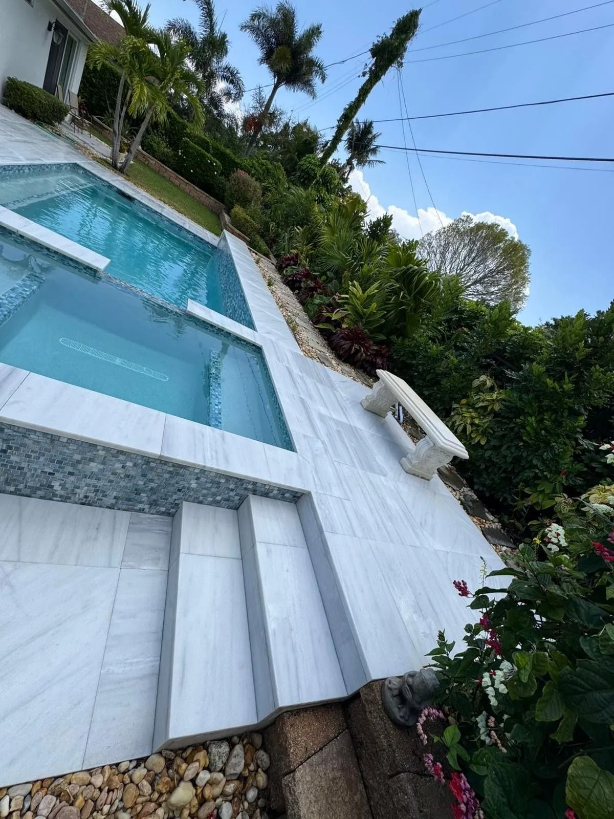 An outdoor pool and spa with white marble tiling, steps, and a bench, surrounded by lush landscaping under a blue sky.