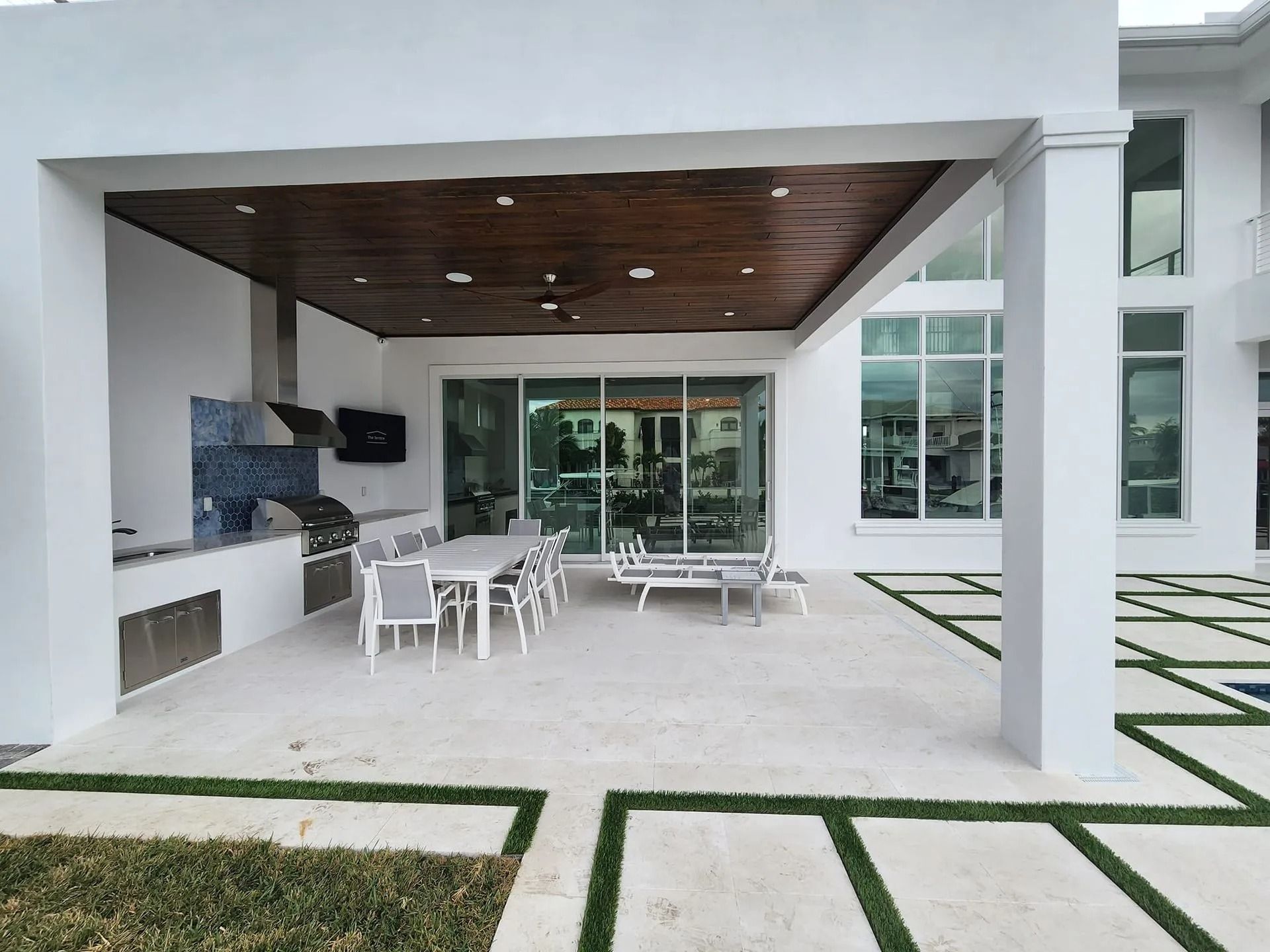 A modern outdoor covered patio with a built-in kitchen, dining area, and large white stone pavers with grass inlays.