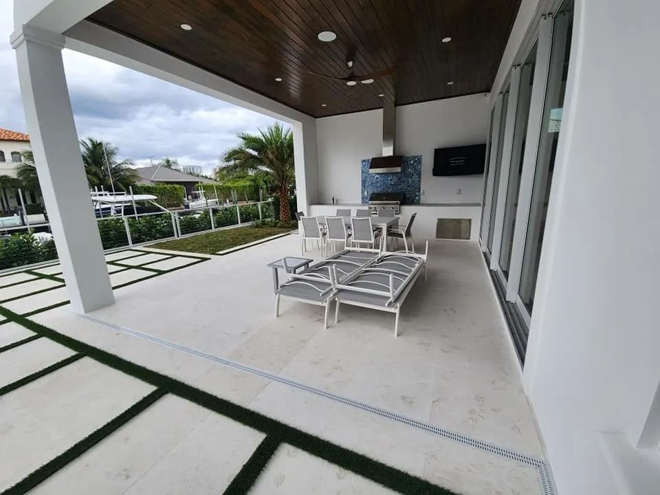 Covered outdoor patio with light stone flooring, white furniture, a built-in kitchen, and views of a lawn and waterway.
