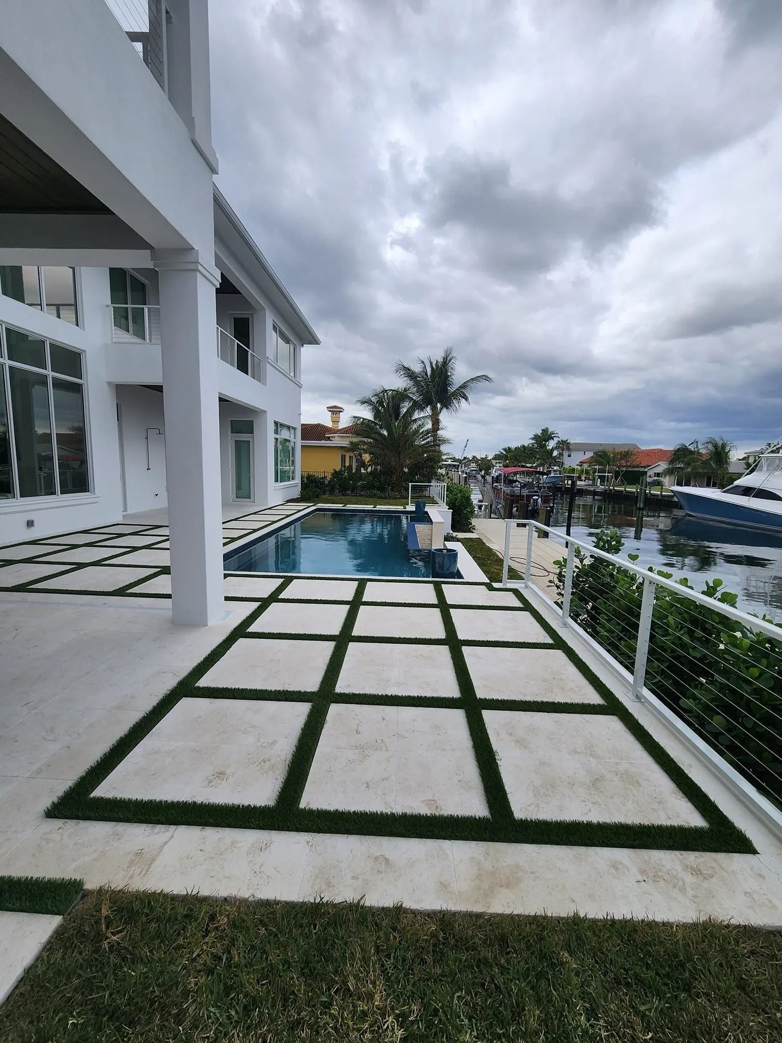 A white multi-story house with a pool and a patio featuring square stone tiles separated by turf borders by the water.
