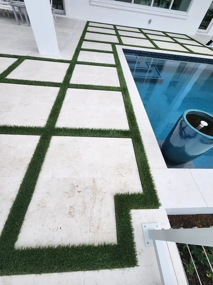 A pool deck featuring square stone pavers separated by strips of green grass, bordering a blue swimming pool.