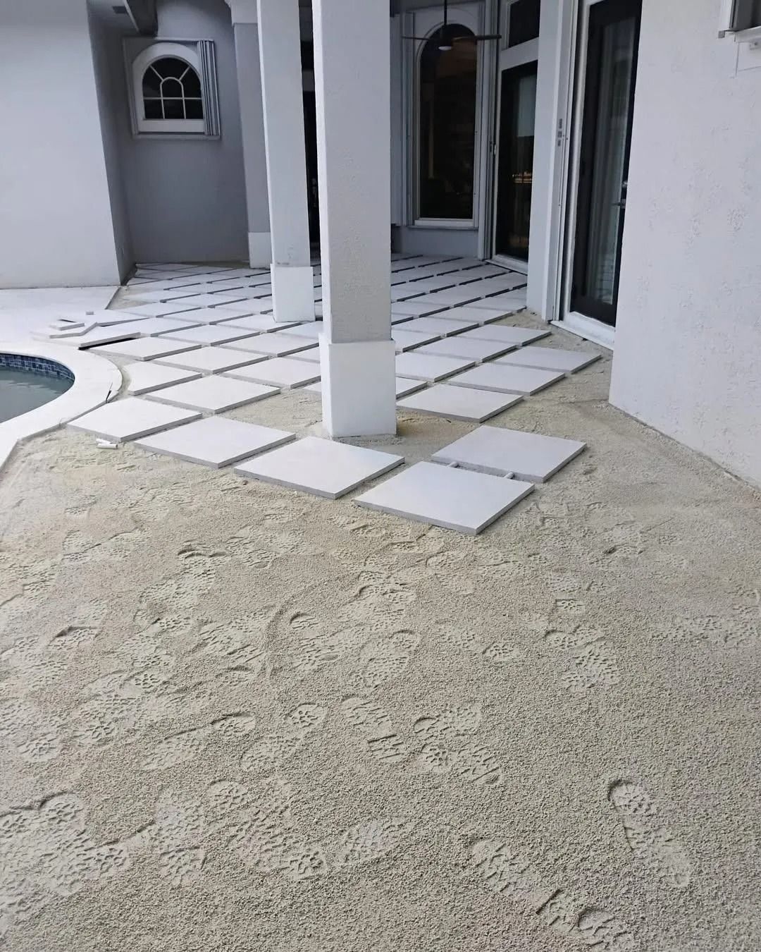 A light-colored patio area with square white pavers set into textured concrete flooring near a pool and house exterior.