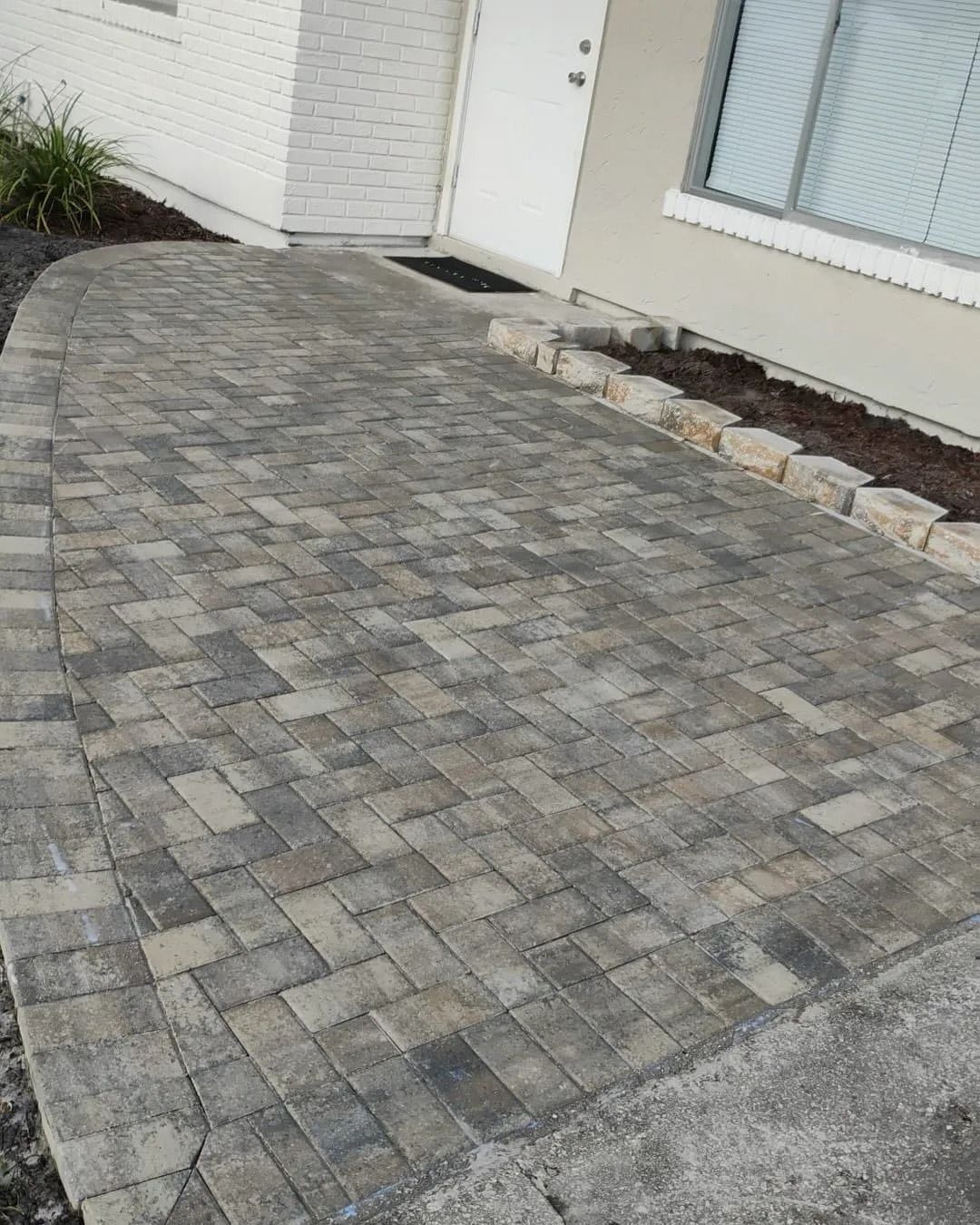 A newly installed grey paver walkway leading to a house entrance with a stone border along the adjacent garden bed.