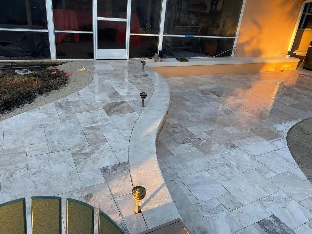 Light-colored stone patio with a curved edge and integrated low-voltage lighting, adjacent to a home with glass doors.
