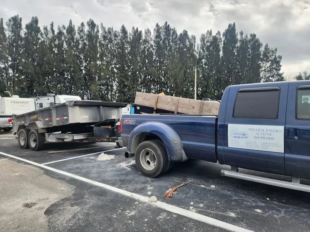 A dark blue pickup truck with a white sign on the door, hitched to an empty silver dump trailer in a parking lot.
