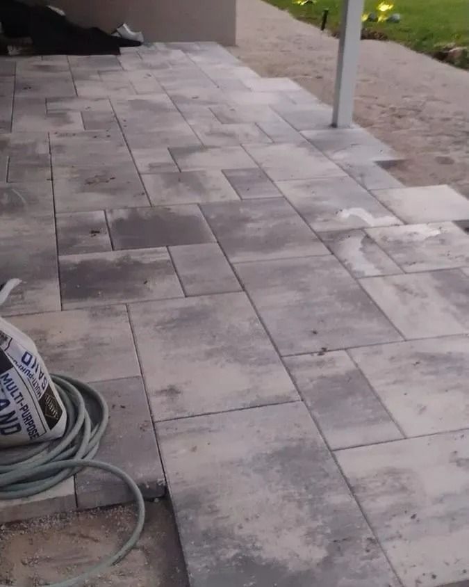 A partially paved outdoor patio features rectangular, grey-toned stone pavers, with a bag of sand and a coiled hose nearby.