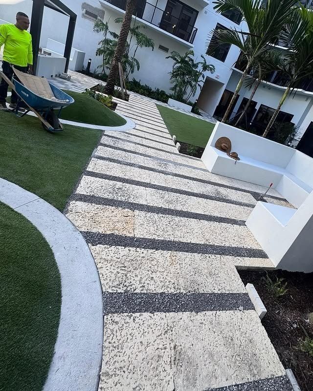 A person in a bright yellow shirt pushes a wheelbarrow along a paved walkway made of stone tiles and dark border accents.