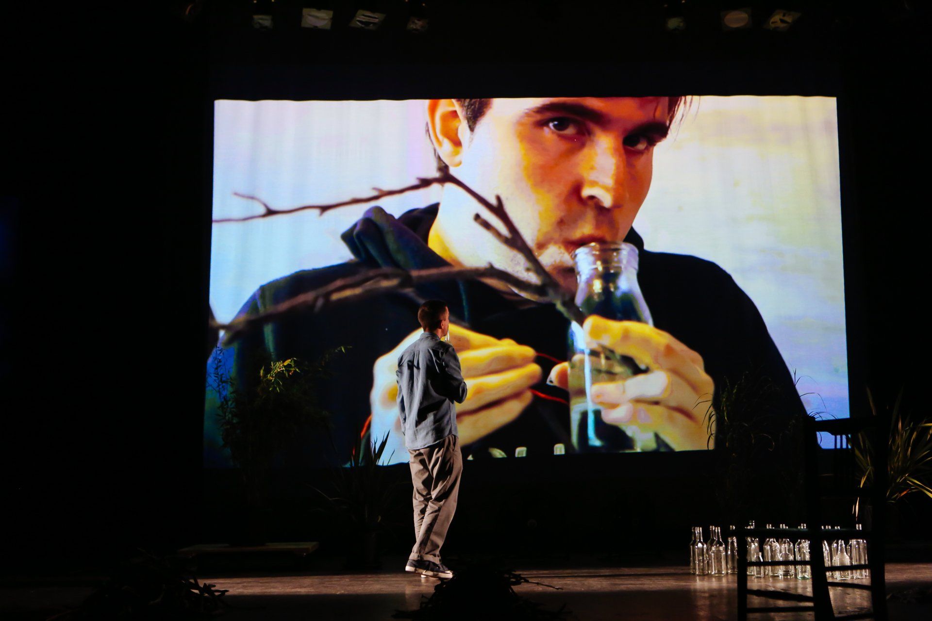 a man is standing in front of a large screen that shows a man drinking from a glass