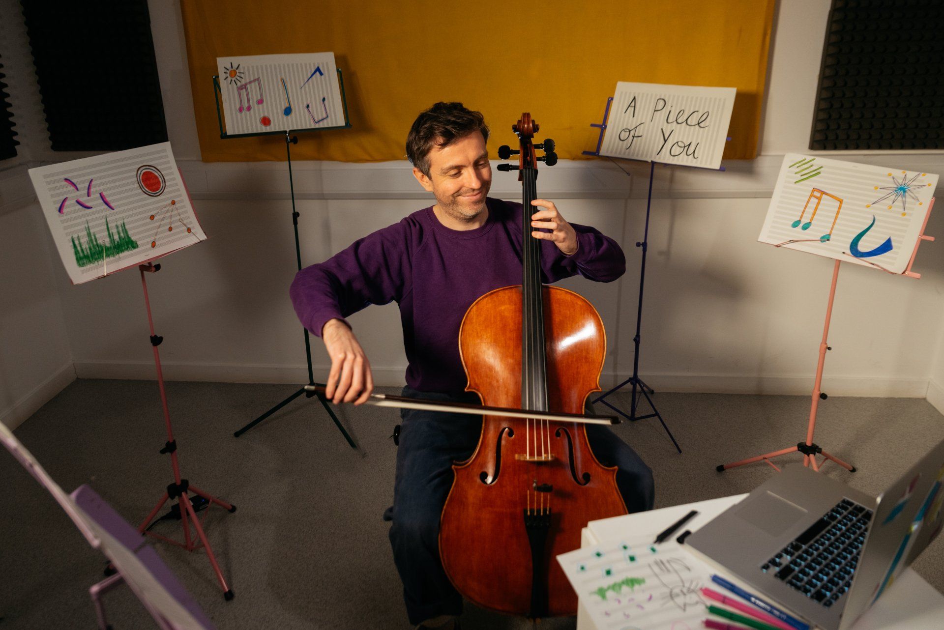 a man is playing a cello in a room with music stands and a laptop .