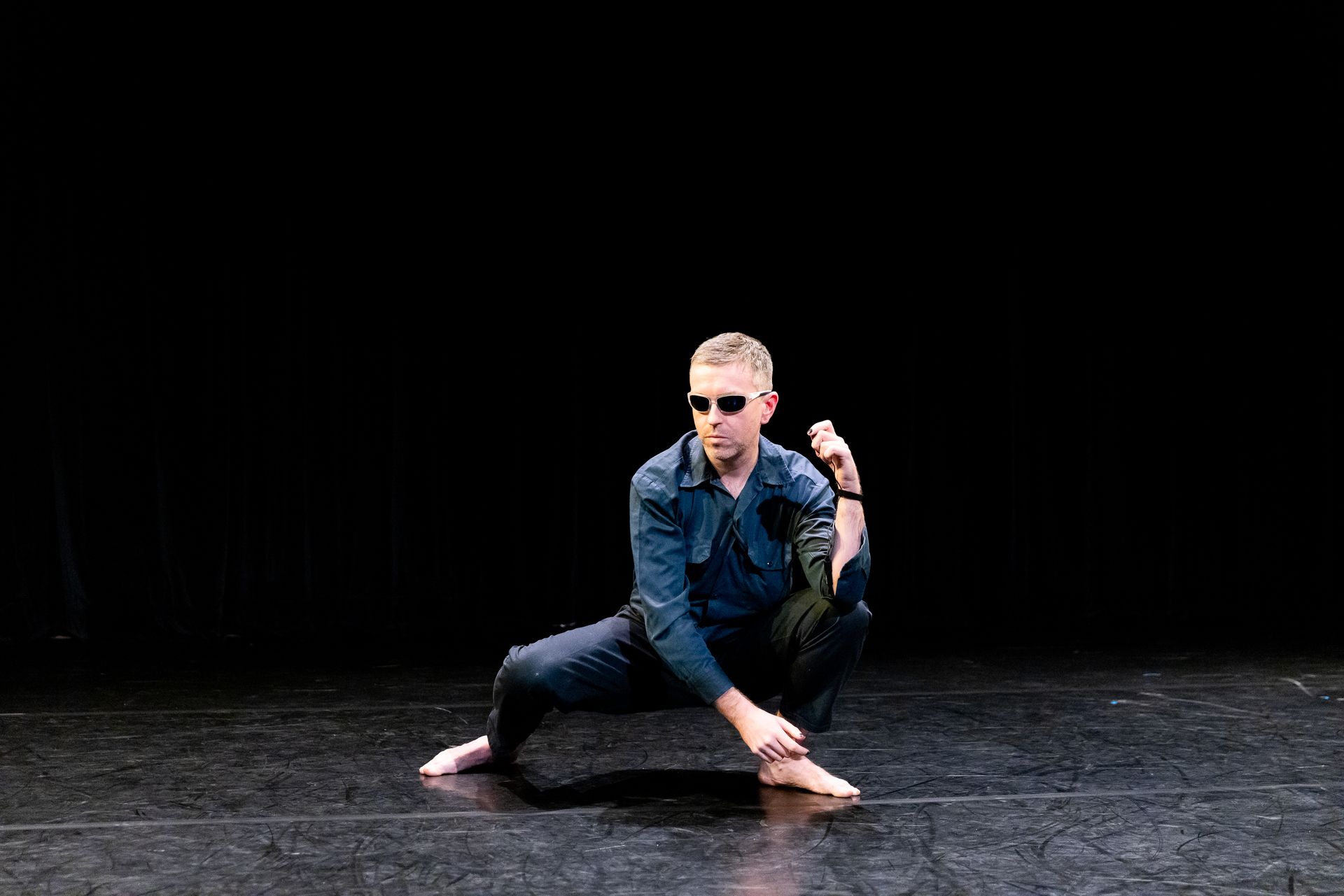 A man wearing sunglasses is squatting on a stage.