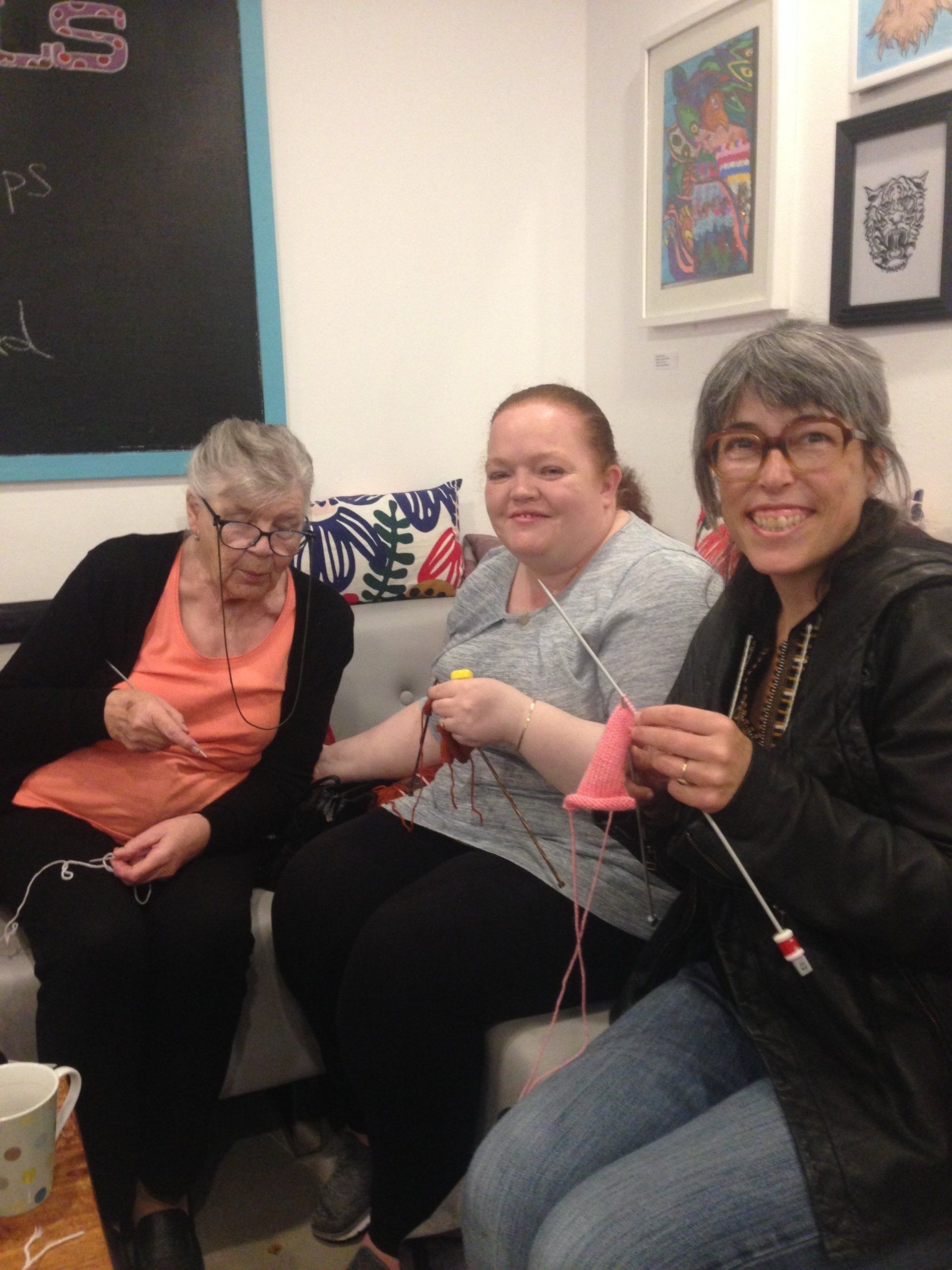three women are sitting on a couch knitting and smiling
