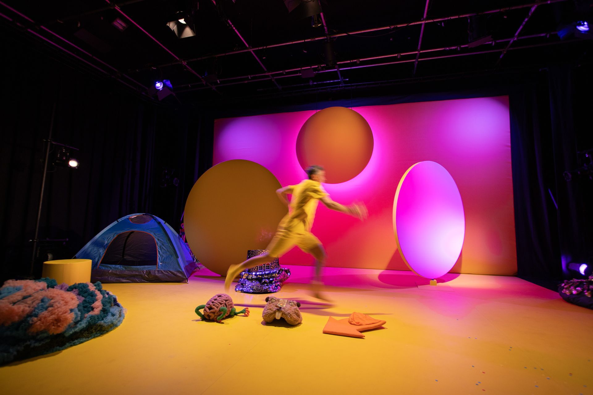 A man in a yellow bodysuit is running on a stage in front of a tent.