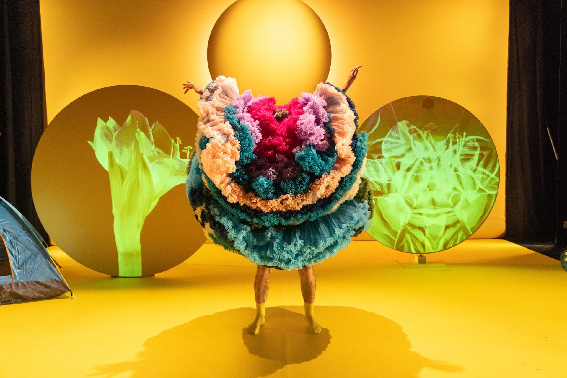 A performer in a colourful dress of layers is standing on a yellow stage.