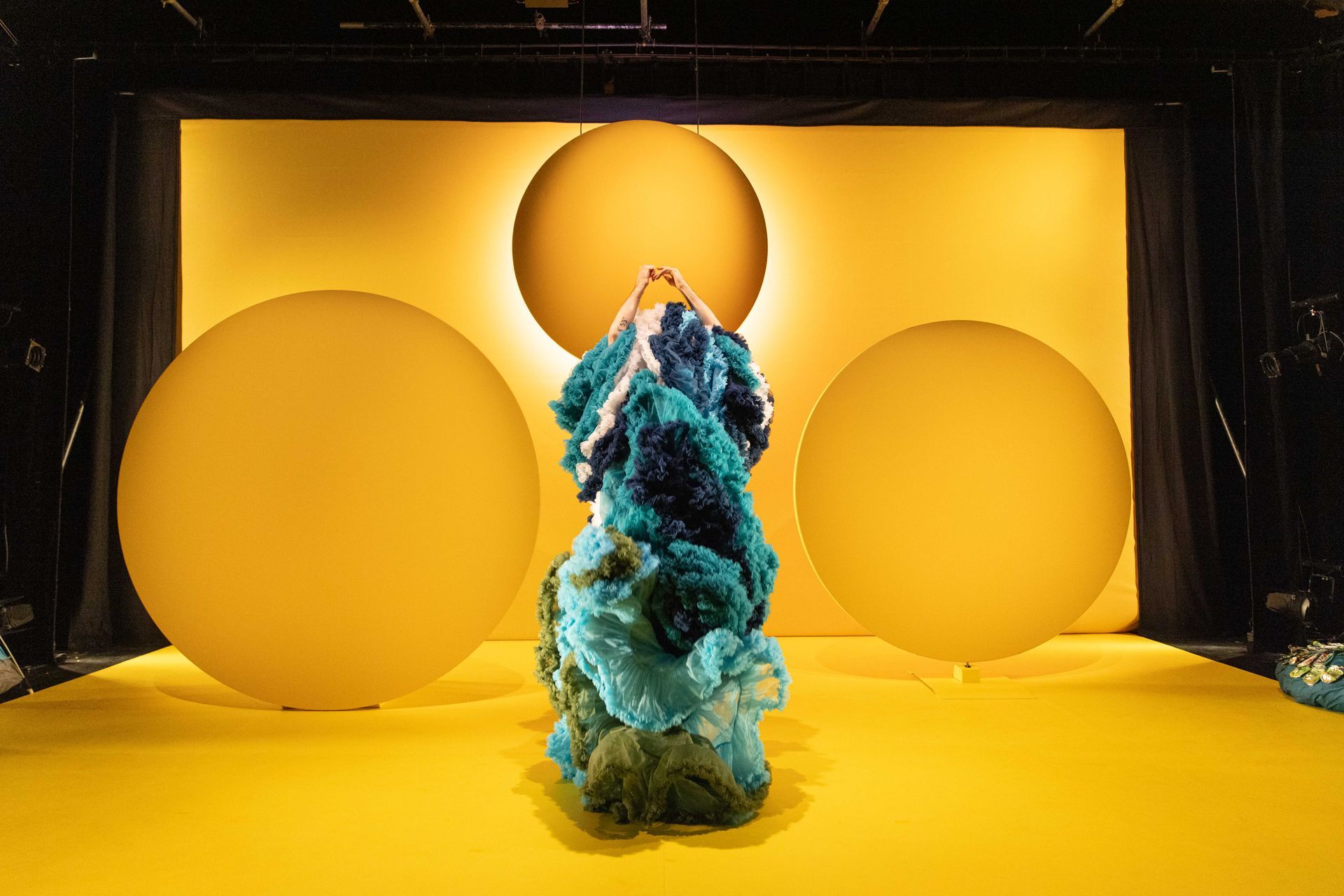 A performer is holding up a column of colourful petticoats in front of a yellow wall.
