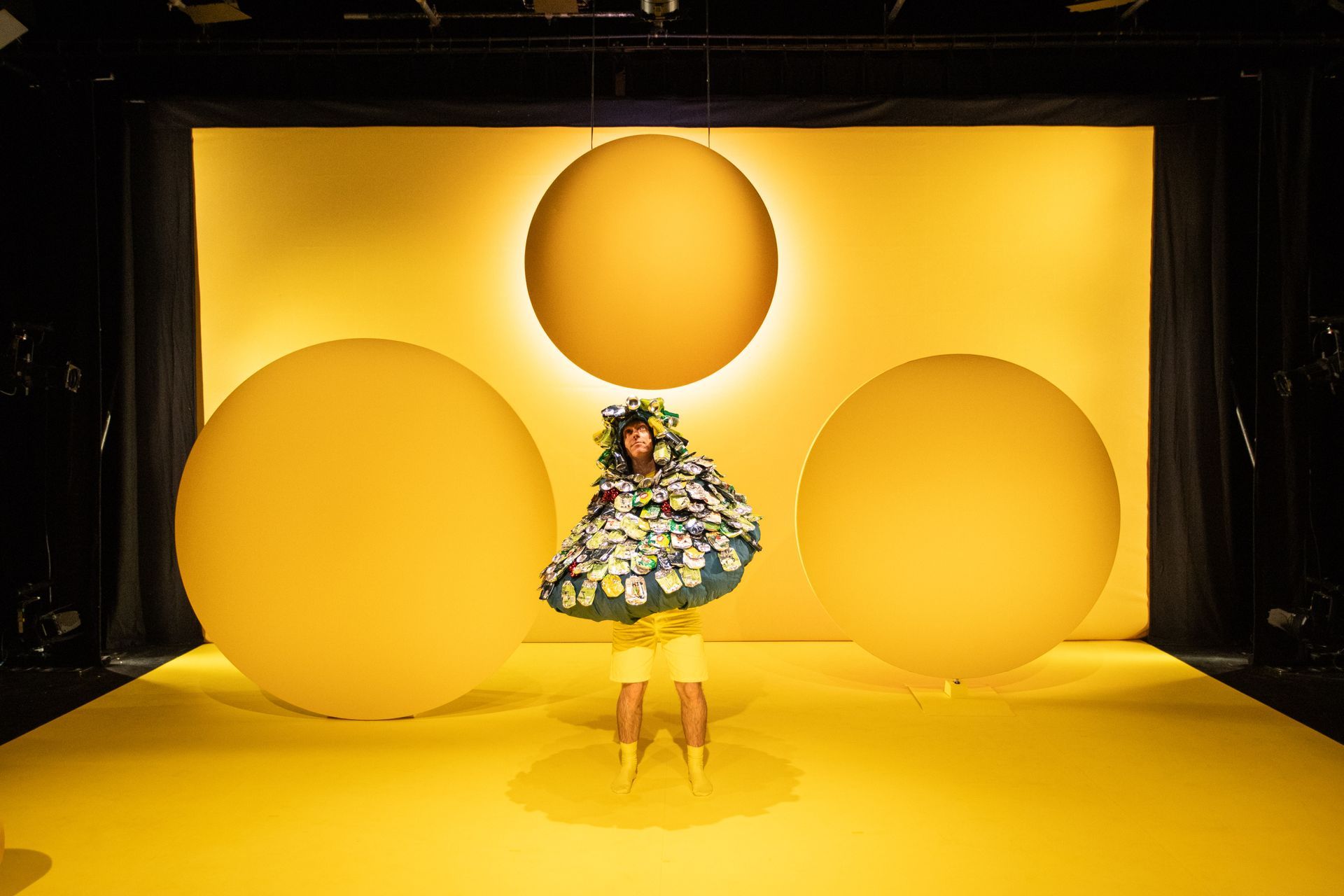 A man in a bulbous green costume covered in crushed cans is standing on a yellow stage.