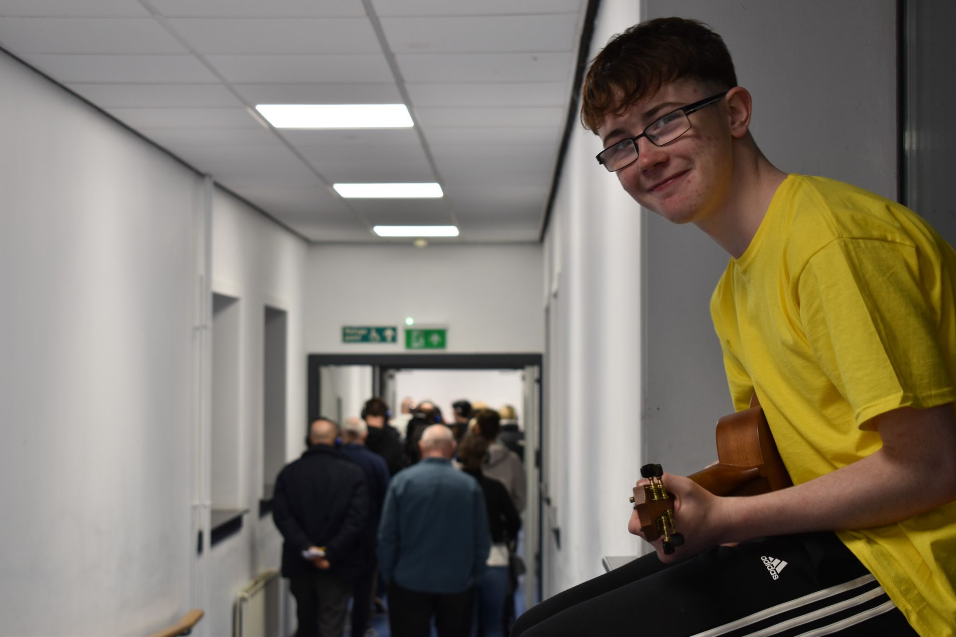 A young man in a yellow shirt is holding a ukulele in a hallway.