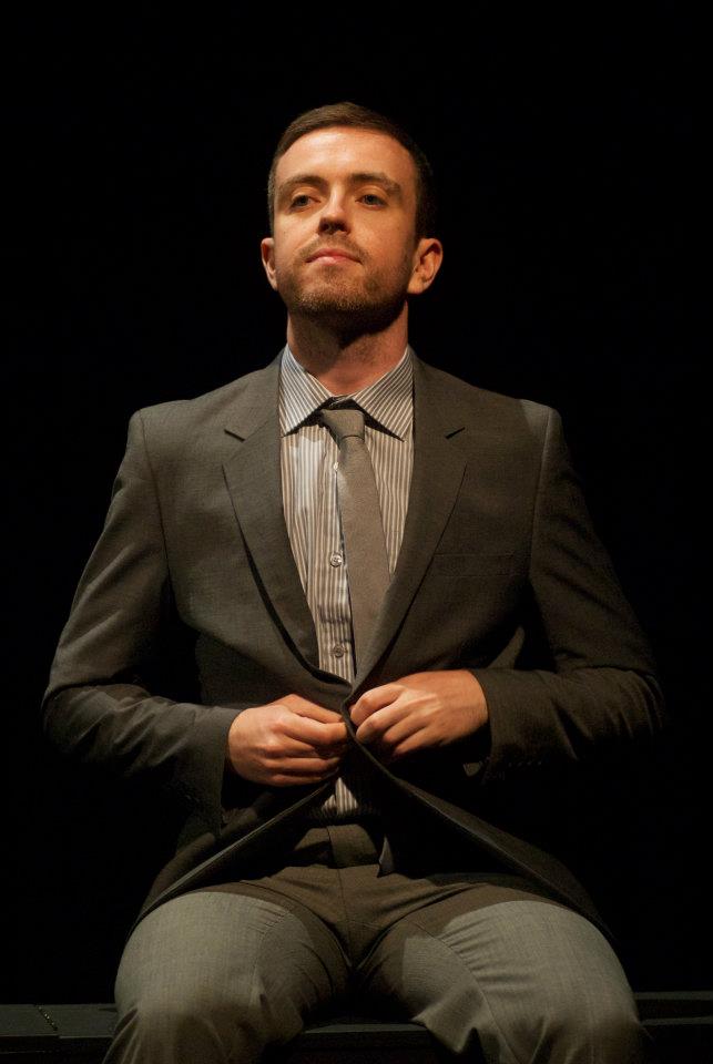 A man in a suit and tie is sitting down and adjusting his jacket.