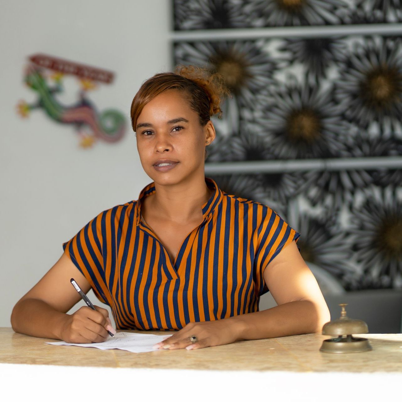 Mujer escribiendo en un escritorio con una campana, una camisa a rayas y obras de arte en el fondo.