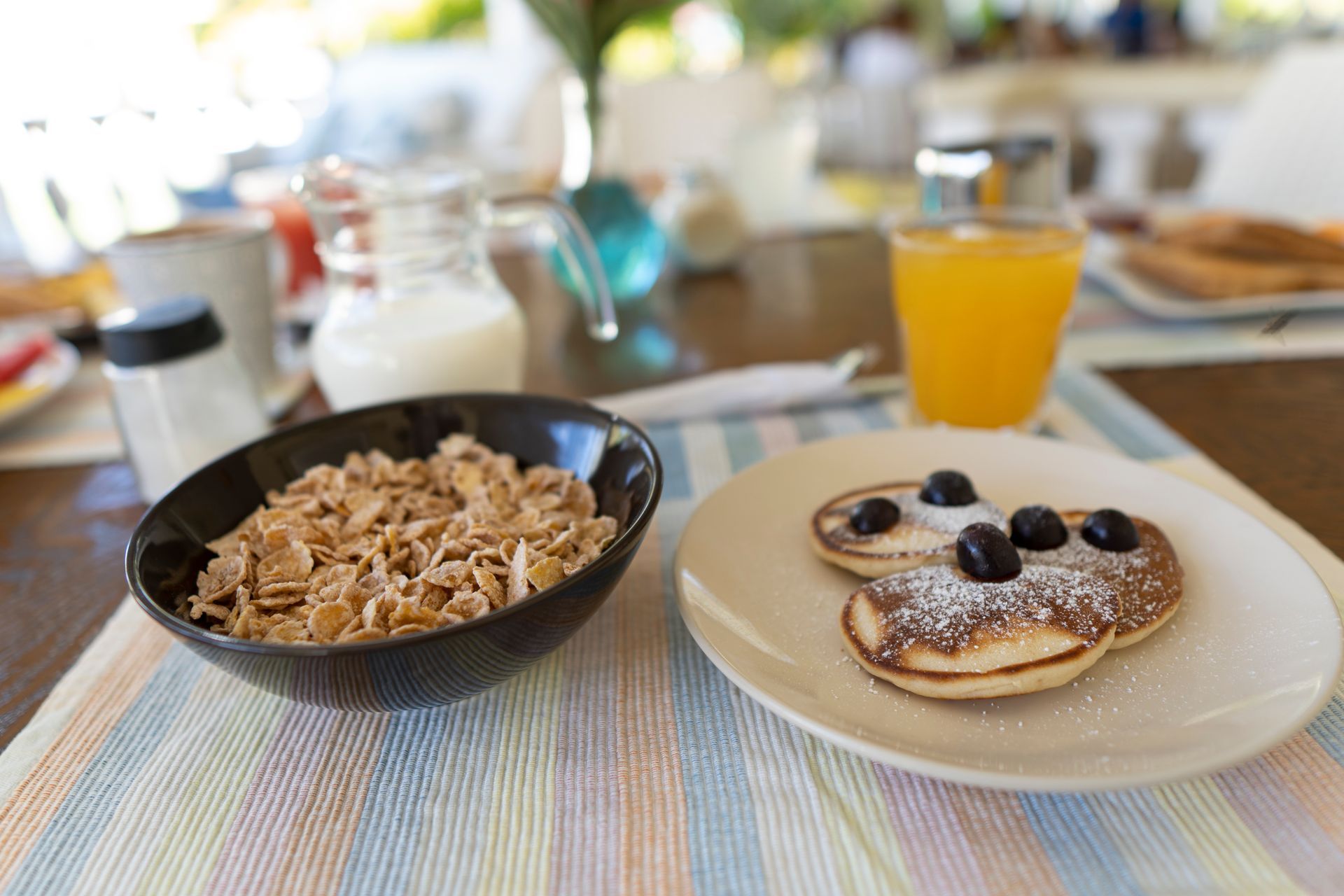 Mesa de desayuno con cereal, panqueques con arándanos, jugo de naranja y condimentos.