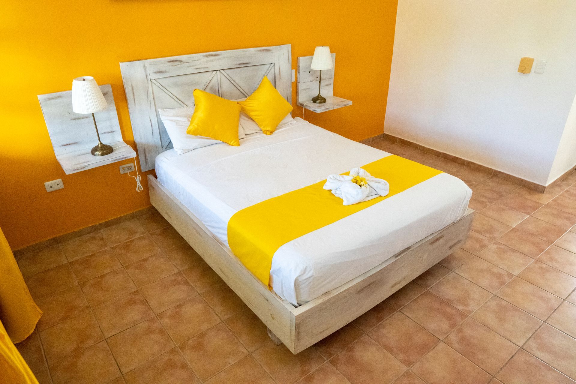 Dormitorio de paredes amarillas con cama, detalles en amarillo, mesitas de noche y suelo de baldosas.
