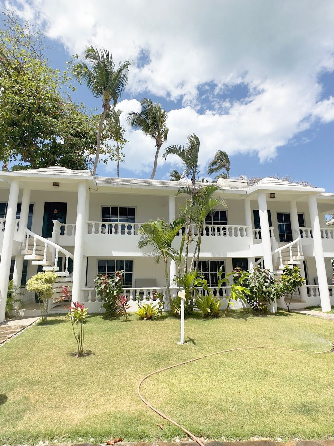 Edificio blanco de dos pisos con balcones, ventanas y palmeras contra un cielo azul.