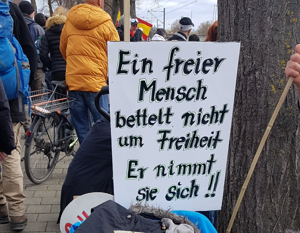 Auf einem Protestschild steht: „Ein freier Mensch bettelt nicht um Freiheit, er nimmt sie sich.“ In einer Menschenmenge im Freien.