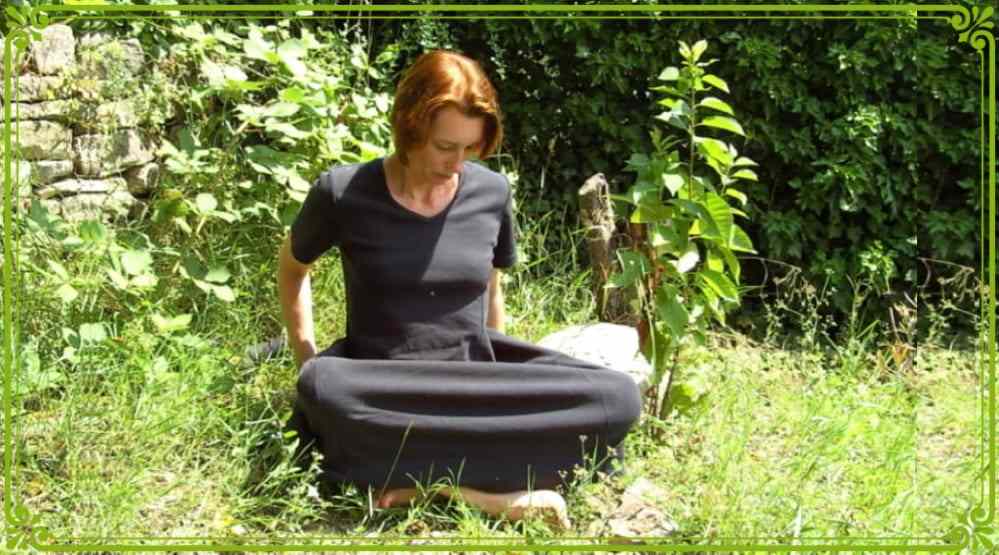 Woman in black dress sitting cross-legged in grassy garden, looking down.
