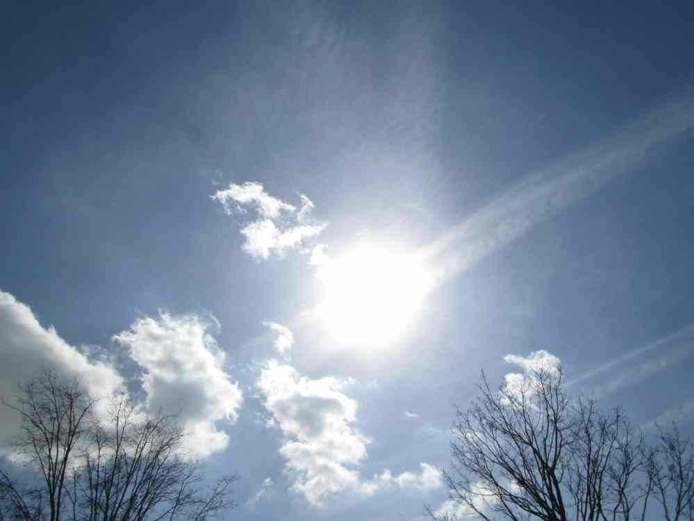 Strahlender Sonnenschein am blauen Himmel, teilweise bewölkt, am unteren Rand zeichnen sich kahle Baumzweige als Silhouetten ab.