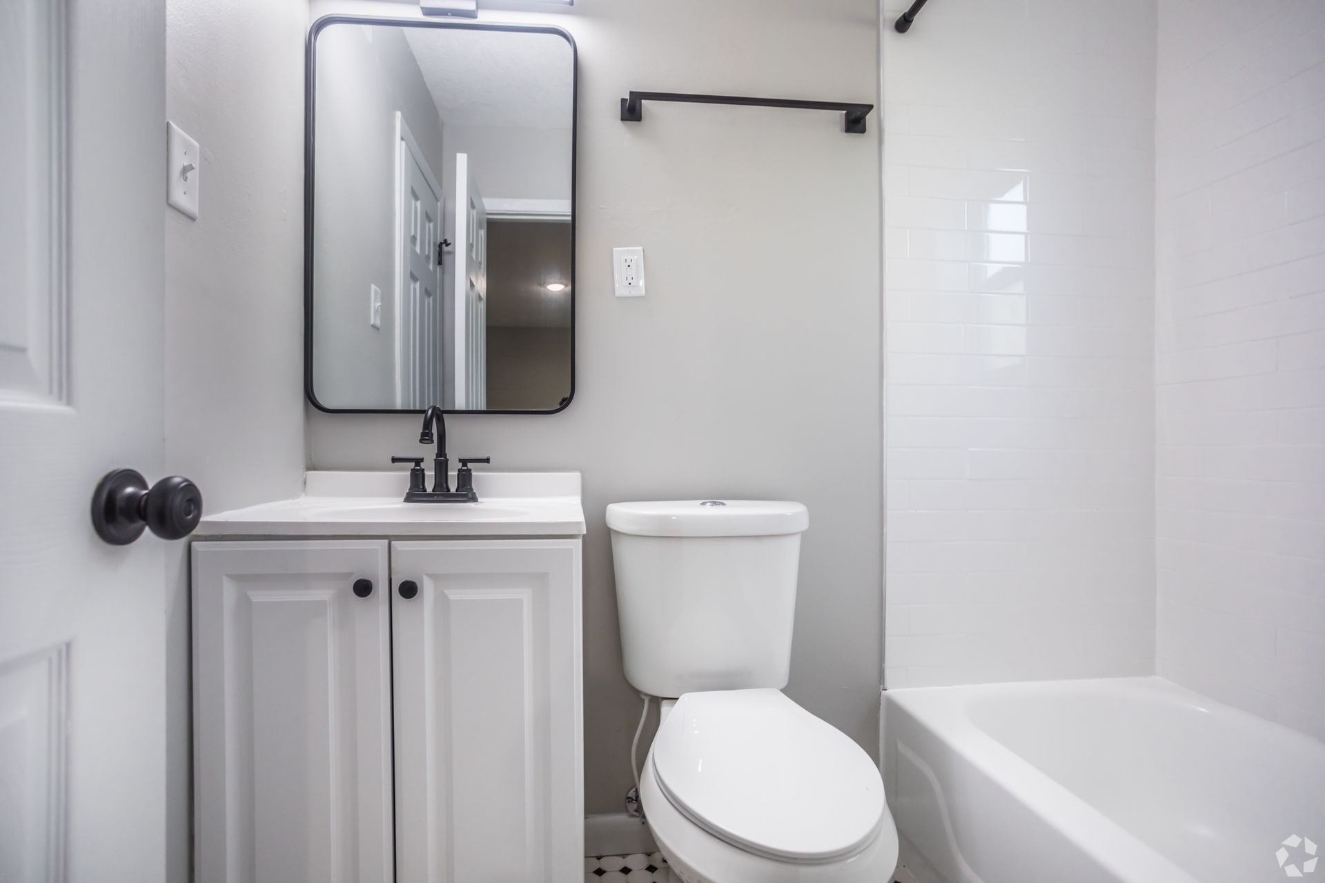 Small, modern bathroom with white walls, vanity, toilet, and bathtub. Black mirror and fixtures.