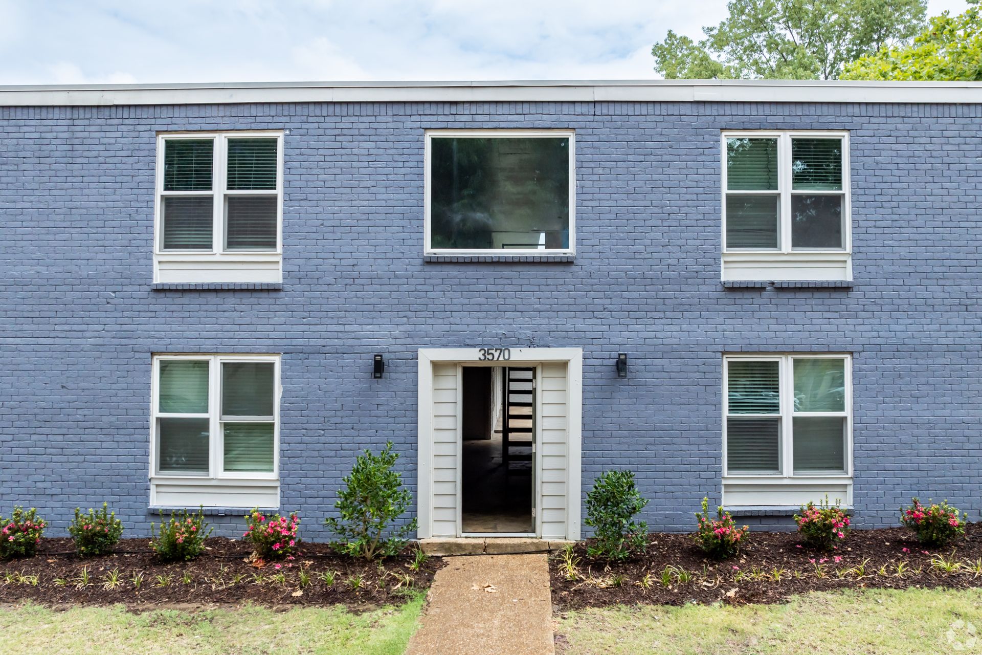 Blue brick building with white-framed windows and an open doorway. Small walkway and landscaping in front.