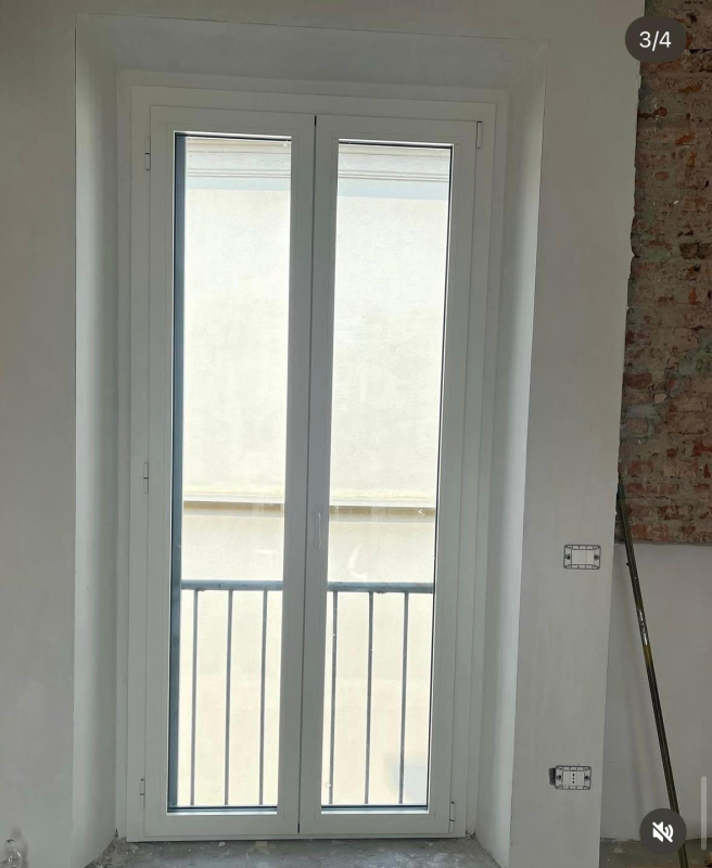 Porte finestre bianche con traverse verticali sui pannelli inferiori, inserite in una parete bianca con una sezione in mattoni a vista sulla destra.