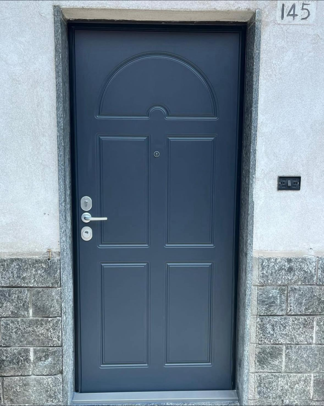 Una porta esterna blu scuro con quattro pannelli rettangolari e una parte superiore arcuata, inserita in una cornice con effetto pietra.