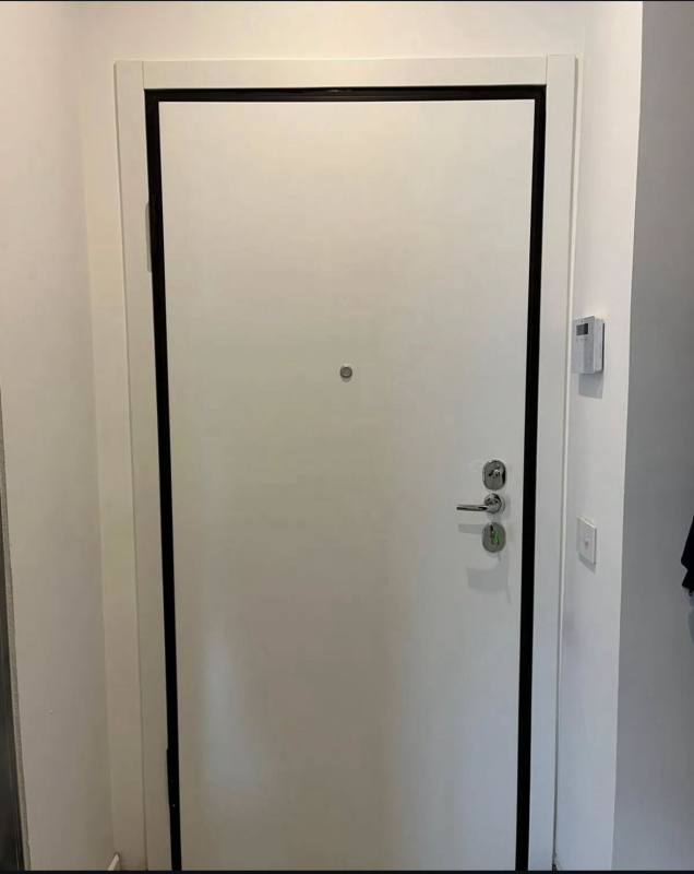 Una porta interna bianca con cornice nera, maniglia argentata, serratura e spioncino, situata in un corridoio bianco.