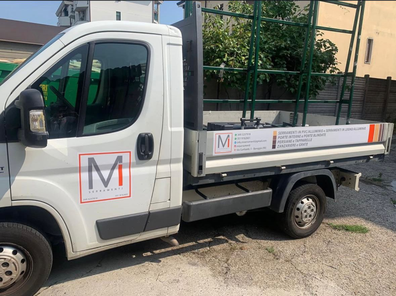 Un camion da lavoro bianco con il logo aziendale raffigurante una grande lettera M e un portapacchi verde per il trasporto del vetro sul pianale.