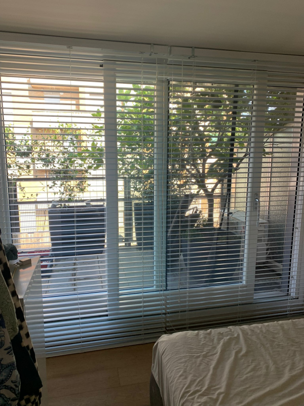 Una vista attraverso persiane orizzontali bianche di un balcone soleggiato con piante in vaso, ripresa da una camera da letto.