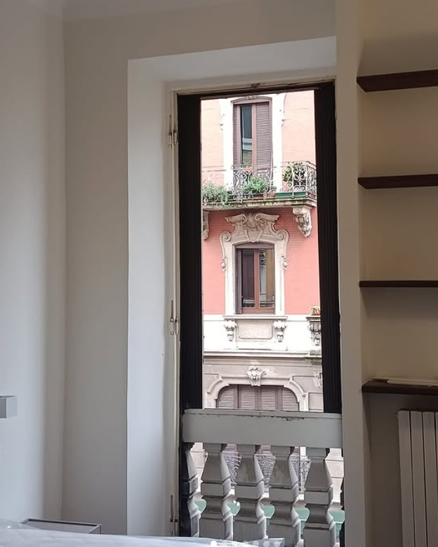 Una vista da una finestra interna che si affaccia su un edificio color salmone con elaborati dettagli architettonici e balconi.