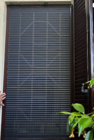 Una mano regge una zanzariera a scomparsa scura e plissettata, installata in un telaio di porta accanto a persiane di legno.