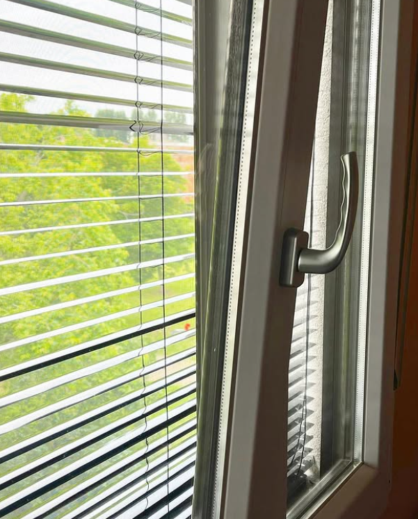 Un telaio bianco della finestra è leggermente aperto, rivelando persiane metalliche argentate e la vista di alberi verdi all'esterno.