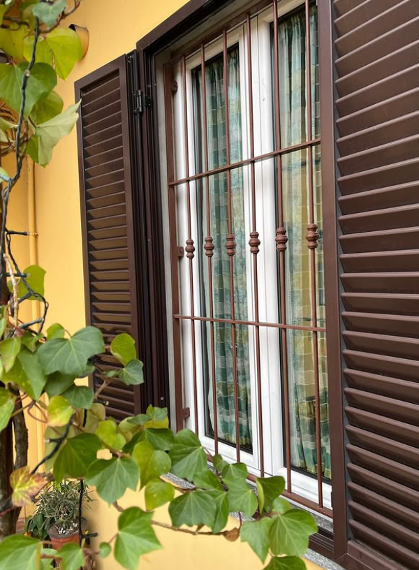 Una finestra con infissi bianchi e sbarre di sicurezza in metallo marrone, affiancata da persiane marroni, accanto a una pianta rampicante verde.